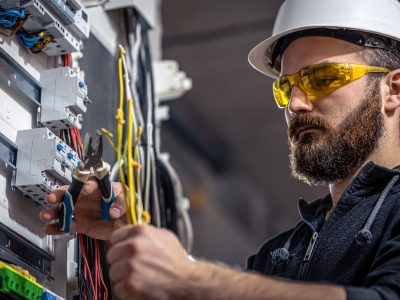 A male electrician works in a switchboard with an electrical connecting cable. Male electrician at the checkout counter on a blurred background of a switchboard.